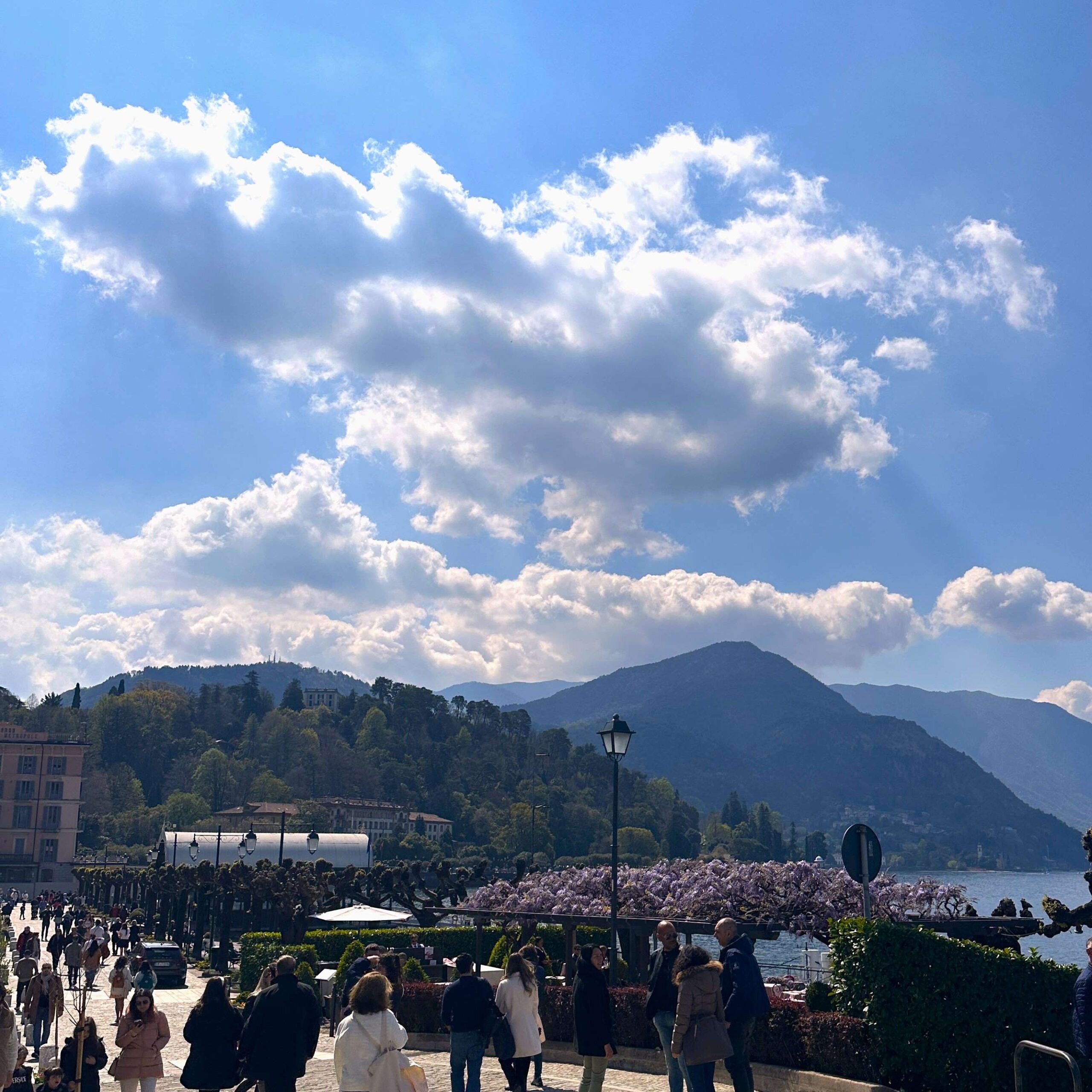Metal red arches hang between a stone wall and the blue water of Lake Como, known as Lovers' Walk. A girl wearing a pink shirt is walking through the archway while taking a photo of the view. In the distance is bright red and yellow buildings. 