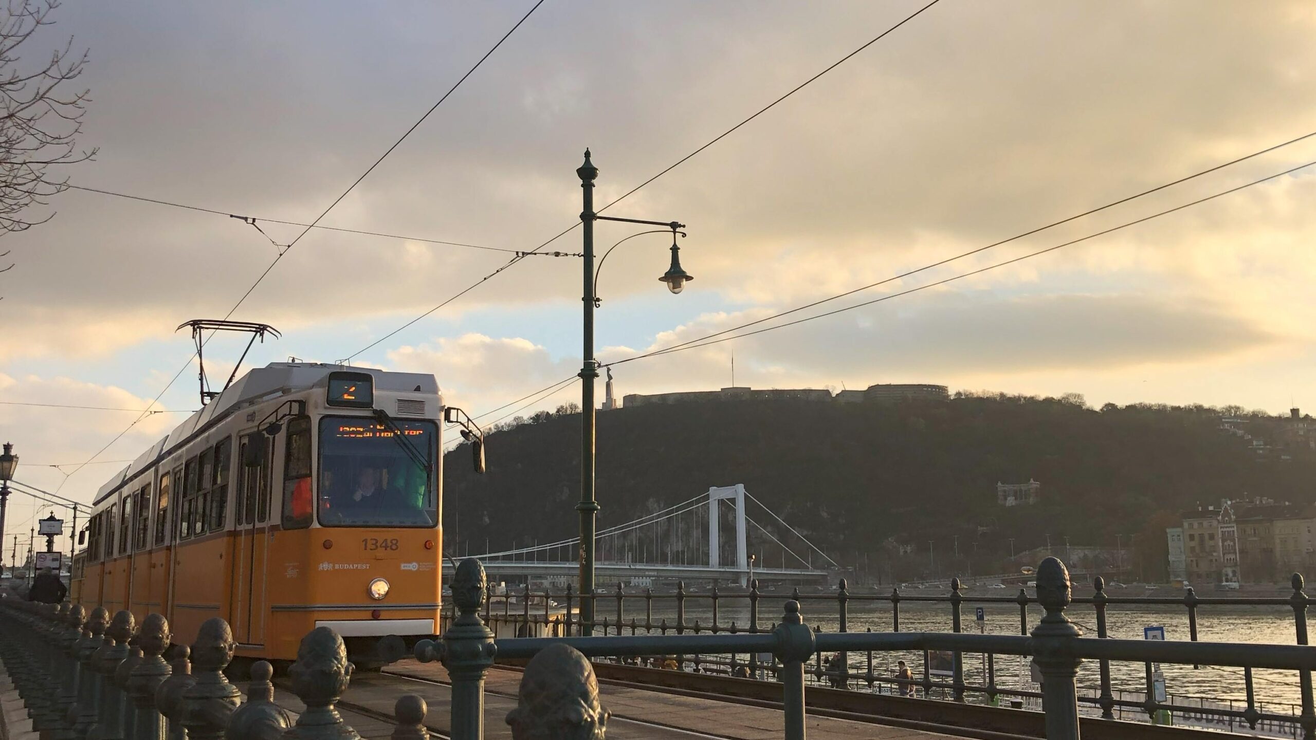 Yellow and white tram pulled up at a tram stop in Budapest. On the outside of the tram, two small stickers with QR codes can be seen next to the door to get on the tram. This is where passengers are requested to scan in order to validate their tram tickets. 