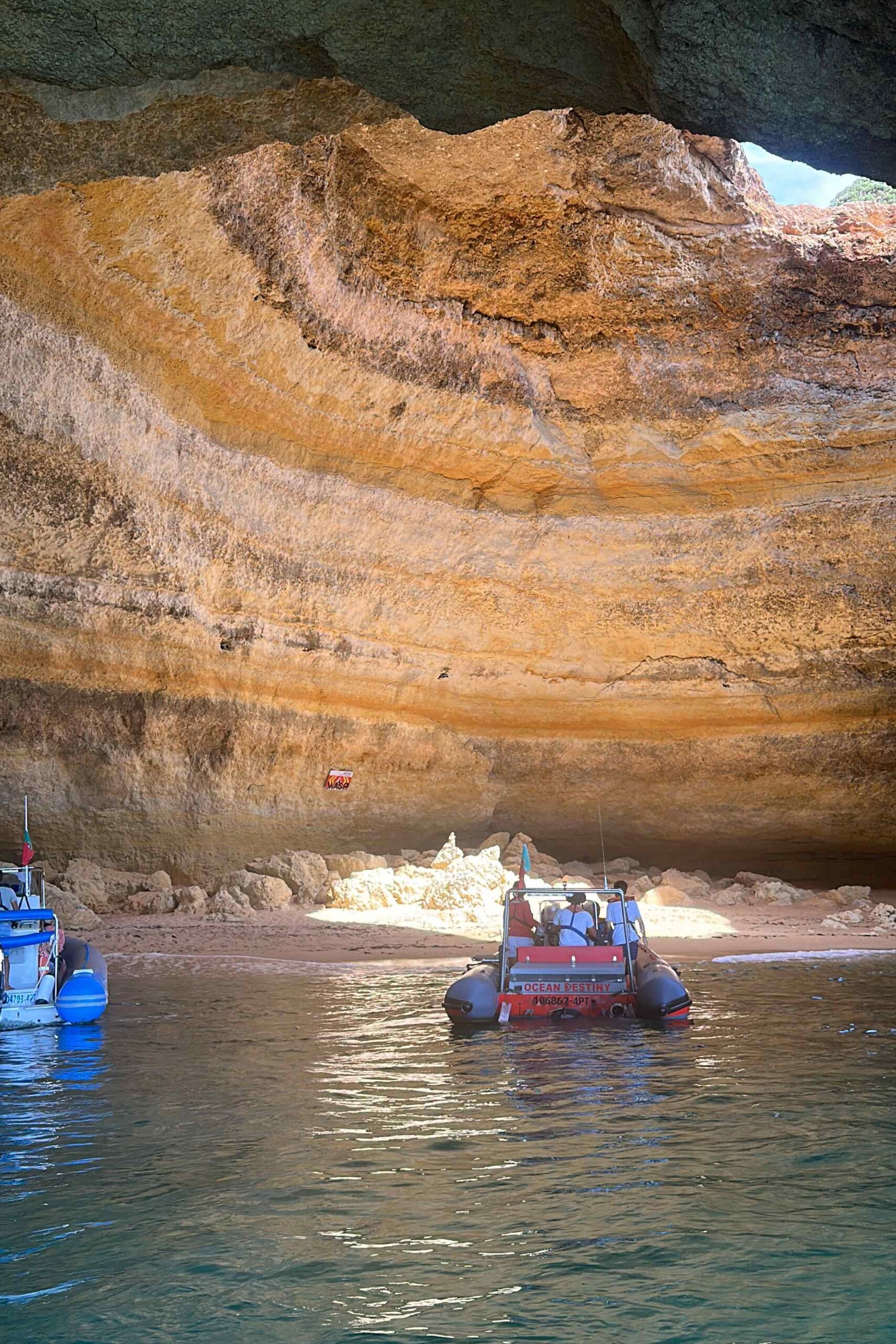Picture of taken from a boat of two other boats going into a cave known as the Benagil Cave. There is a hole in the top of the cave with sunlight beaming through.