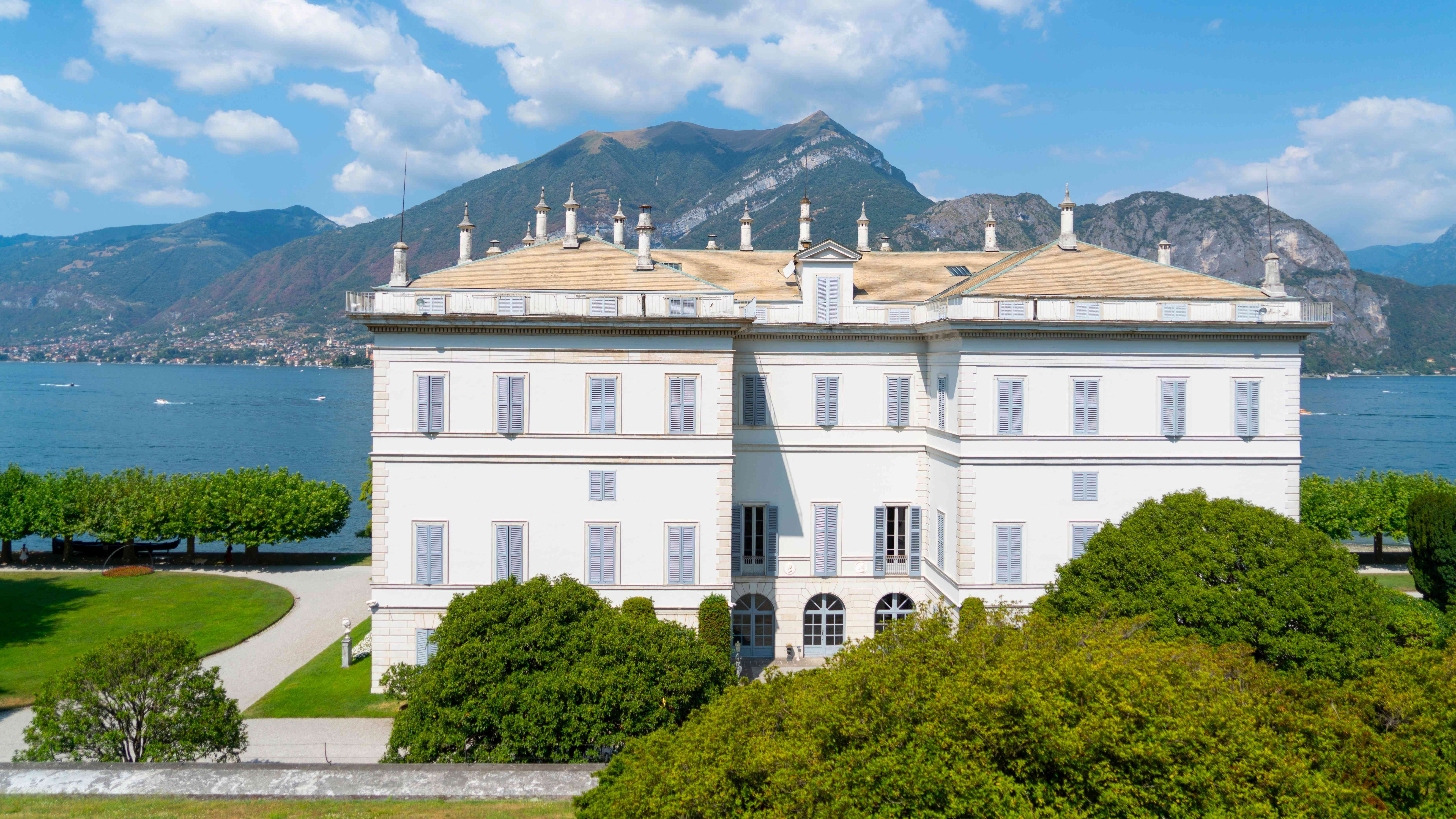 View from the point of view of looking through the trees. Dark green trees cover either corner of the landscape, in the middle is the blue water from Lake Como. Part of a mountain is pictured in the distance, with many white and yellow houses at the bottom of the mountain, overlooking the lake. 